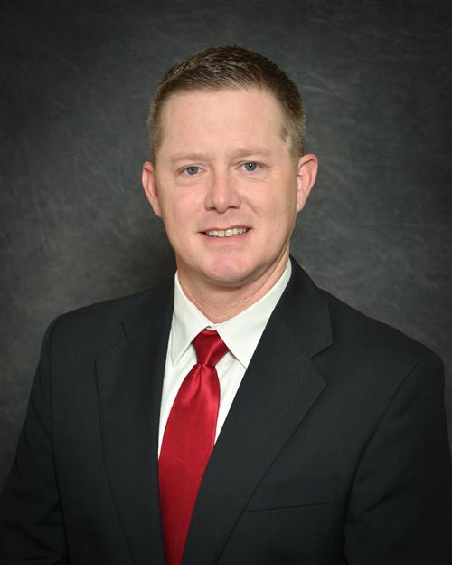 Jonathan Taylor's professional headshot; wearing a black suit and red tie against a grey background