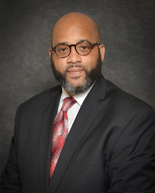 Dr. Brooks' professional headshot; wearing a black suit and red tie against a grey background