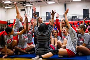 A group of young cheerleaders in grey T-shirts and red shorts is seated on a gym floor, raising their arms and cheering. They
