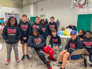 A group of young students wearing matching black hoodies with "PCIS Robotics 2024" printed on them sit together in a school g