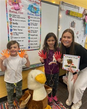 Two young students holding paper cutouts shaped like hands, representing a paper hug. They stand with a teacher who holds a b