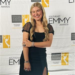  A young woman in an elegant black dress with sheer sleeves smiles brightly at the Southeast Emmy Aw