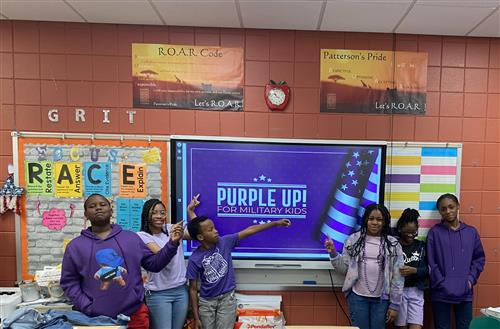 A group of six students standing in front of a digital screen that displays 'Purple Up! For Military Kids.' The students are 
